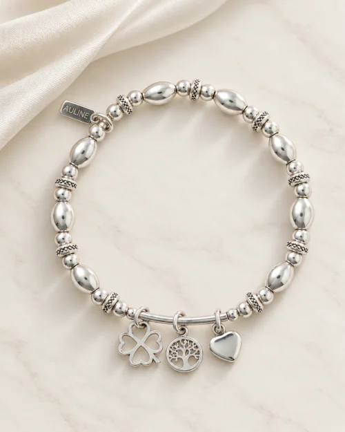 Pulsera Nexo Tri de plata de ley 925 con cuentas facetadas y ovaladas y tres colgantes de trébol, árbol de la vida y corazón, sobre base de mármol color crema